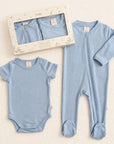 A baby sleepwear set in light blue, featuring a short-sleeve bodysuit, a long-sleeve footed sleeper, and the original window box packaging on a textured cream background.
