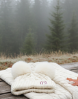 White knitted blanket and white double pom knitted beanie on a wooden bench with a misty forest background