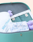 Close-up view inside the teal keepsake trunk showing a personalized white baby blanket embroidered with the name "Matthew" and matching teal apparel.