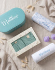 An aesthetic lifestyle flat lay on textured fabric featuring the personalized teal trunk, boxed apparel, and rolled blankets with "Matthew" embroidery.