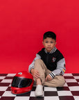 A young boy wear a black and gray Bomber Jacket - Mickey 28 varsity jacket and Native shoes next to a red full-face helmet.