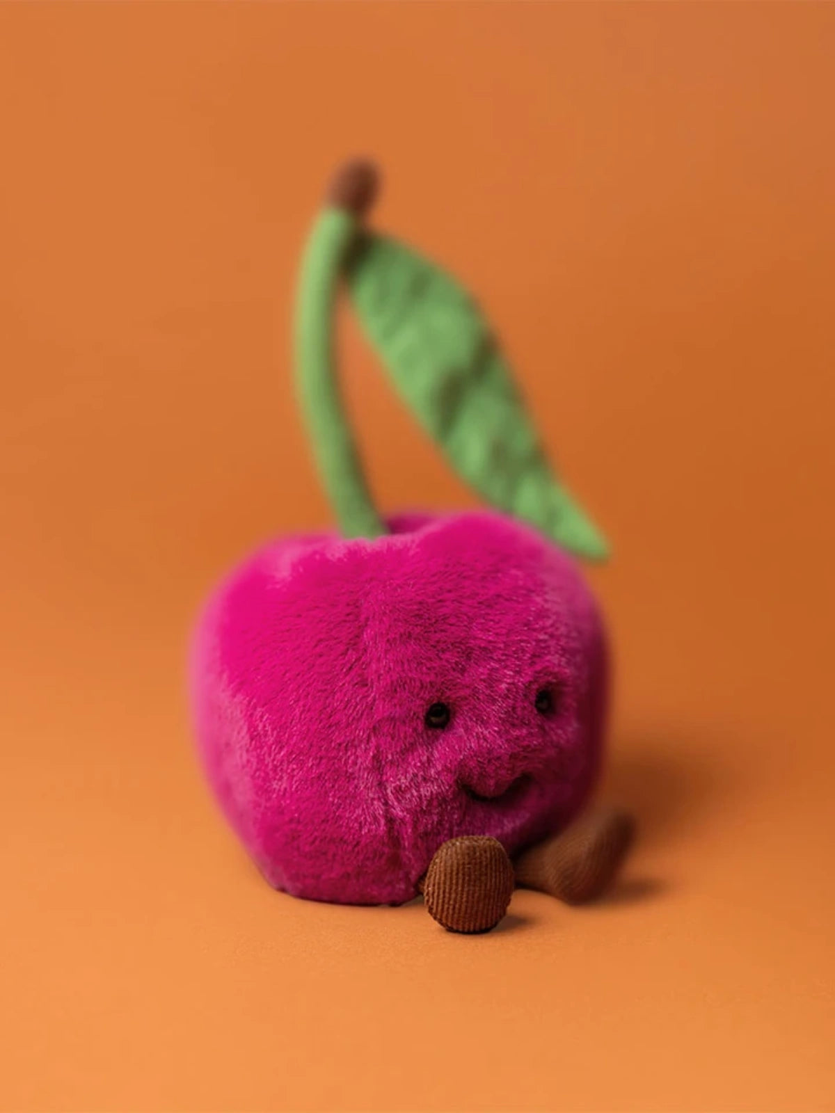 Amuseables Cherry plush toy angled on orange background with smiling face