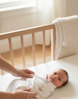 Baby lying in a crib with a person's hand adjusting the swaddle