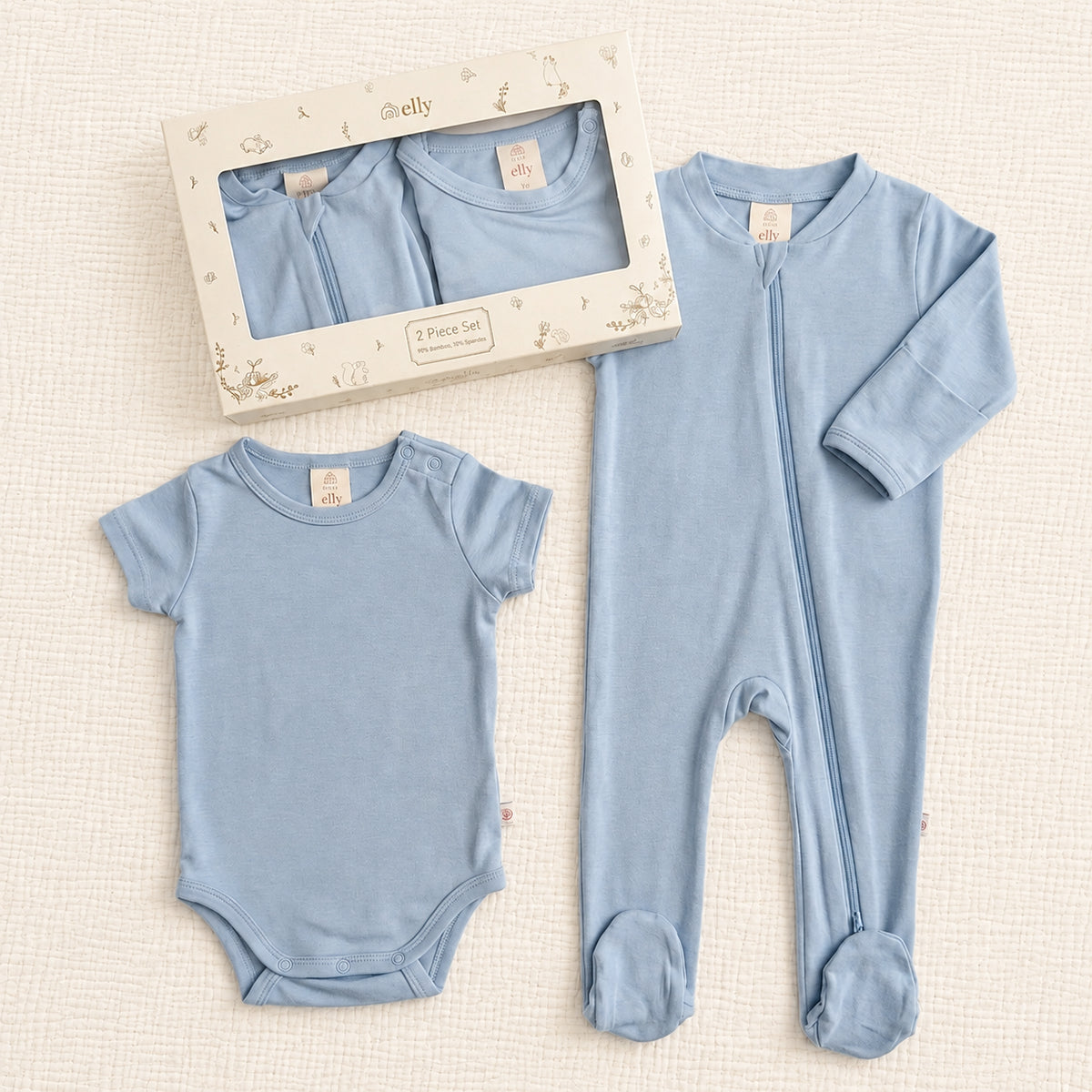 A baby sleepwear set in light blue, featuring a short-sleeve bodysuit, a long-sleeve footed sleeper, and the original window box packaging on a textured cream background.