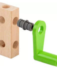 A wooden toy block with three circular holes on one side, connected to a bright green crank handle and a gray plastic fitting, shown on a plain white background.