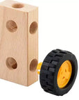 A wooden toy block with pre-drilled holes next to a small black tire-shaped wheel with a yellow center, shown on a plain white background.