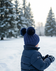 Child wearing a blue winter coat and blue knit beanie with double pom-poms 