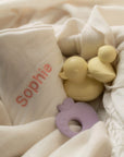 Yellow duck and purple teether on a white blanket with 'sophie' embroidered.