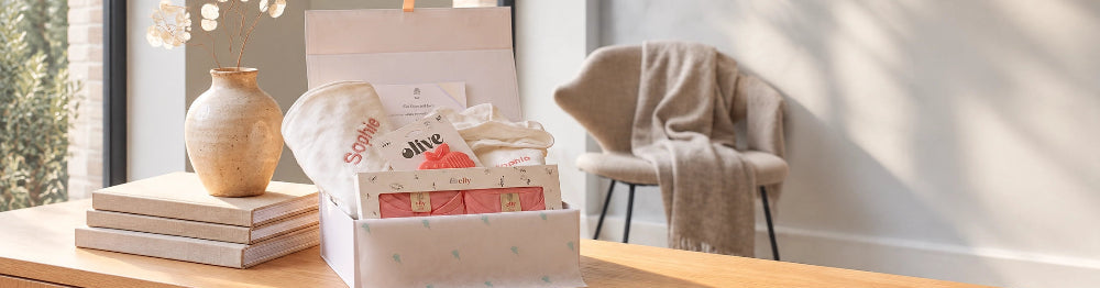 A curated gift box filled with baby essentials, including a white knit hat and packaged items, displayed on a wooden table in a bright, modern room with a chair and window.