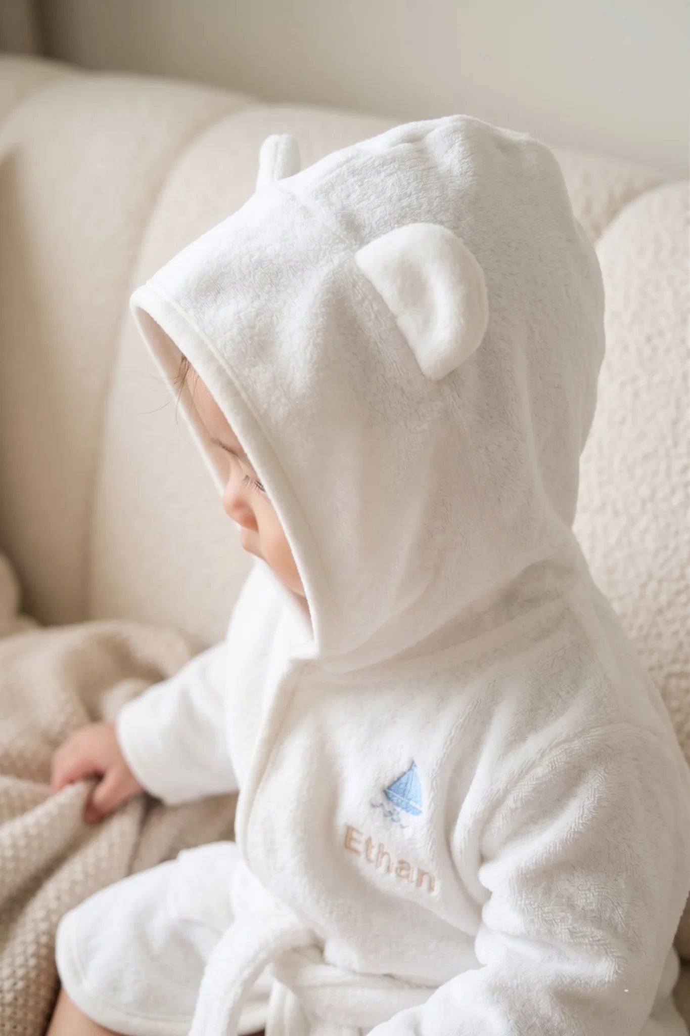 Side view of baby in white hooded bear bathrobe showing embroidery and soft texture