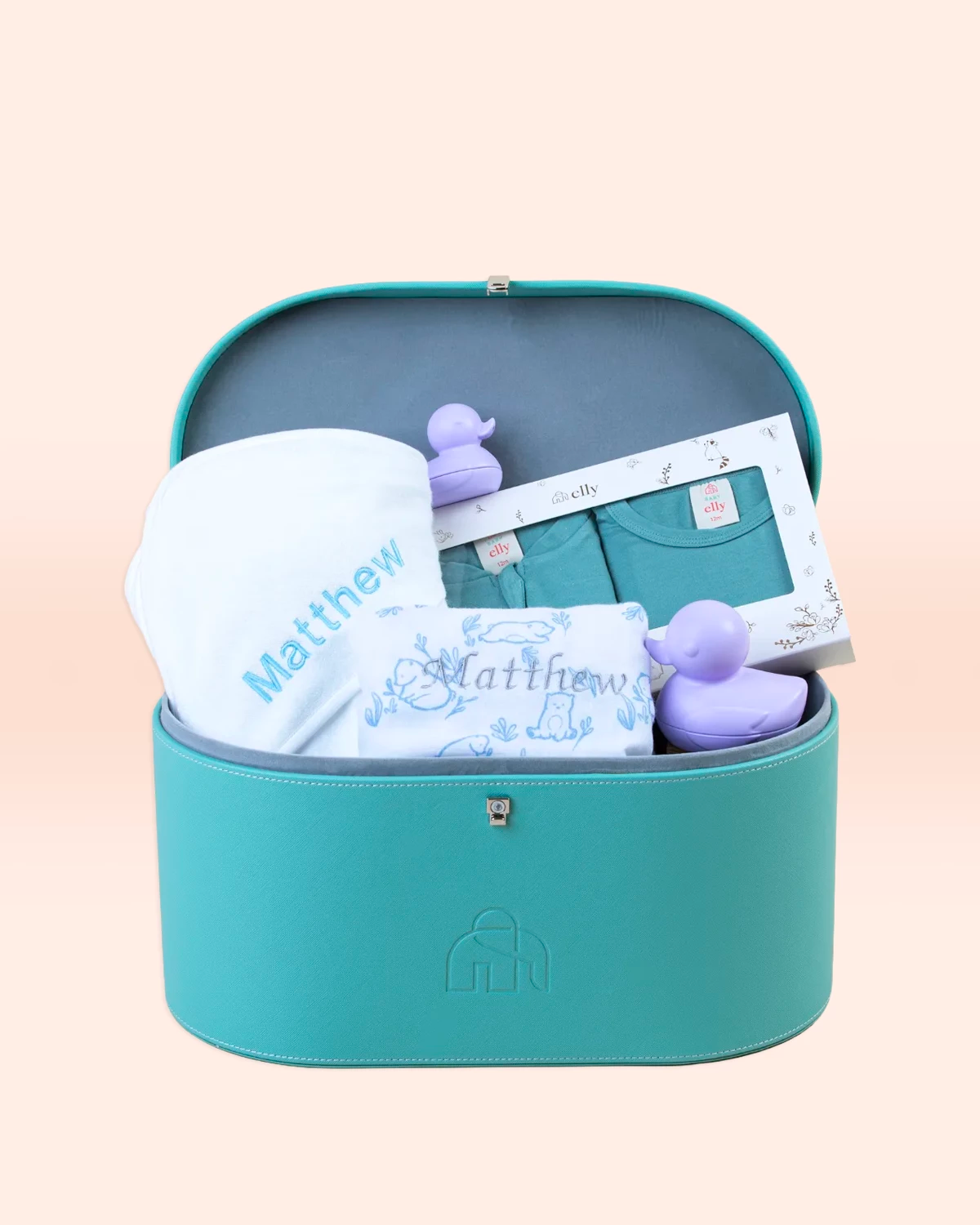 A teal vegan leather keepsake trunk open to reveal a personalized baby gift set including a white blanket, teal clothing, and purple rubber ducks.