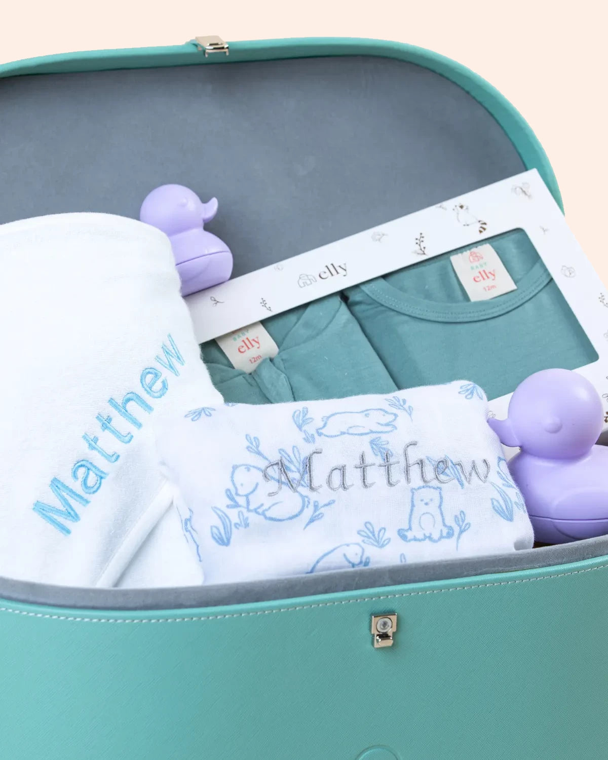 Close-up view inside the teal keepsake trunk showing a personalized white baby blanket embroidered with the name "Matthew" and matching teal apparel.