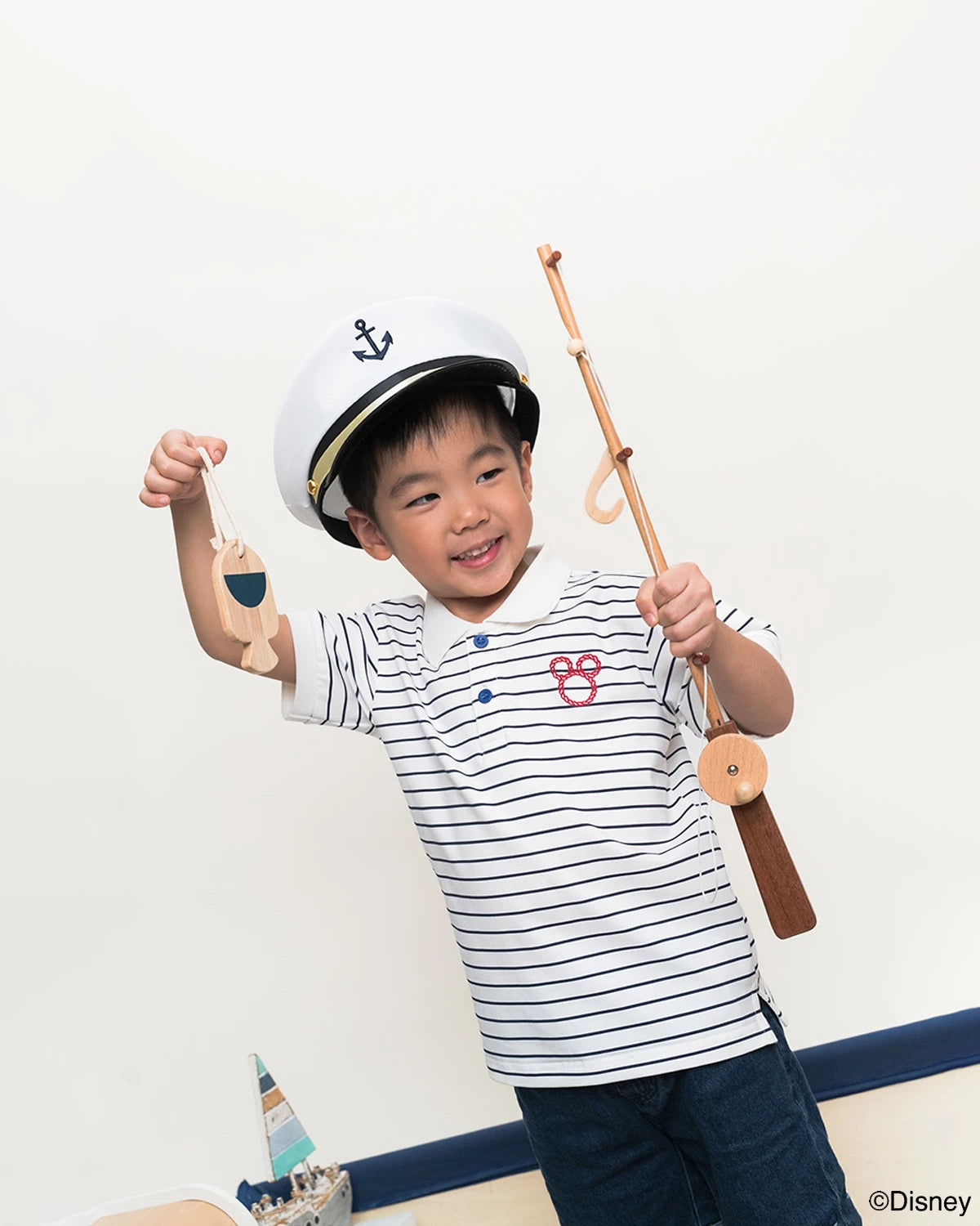 Child wearing a sailor hat and polo tee in stripe sailor mickey holding a toy boat and oar.