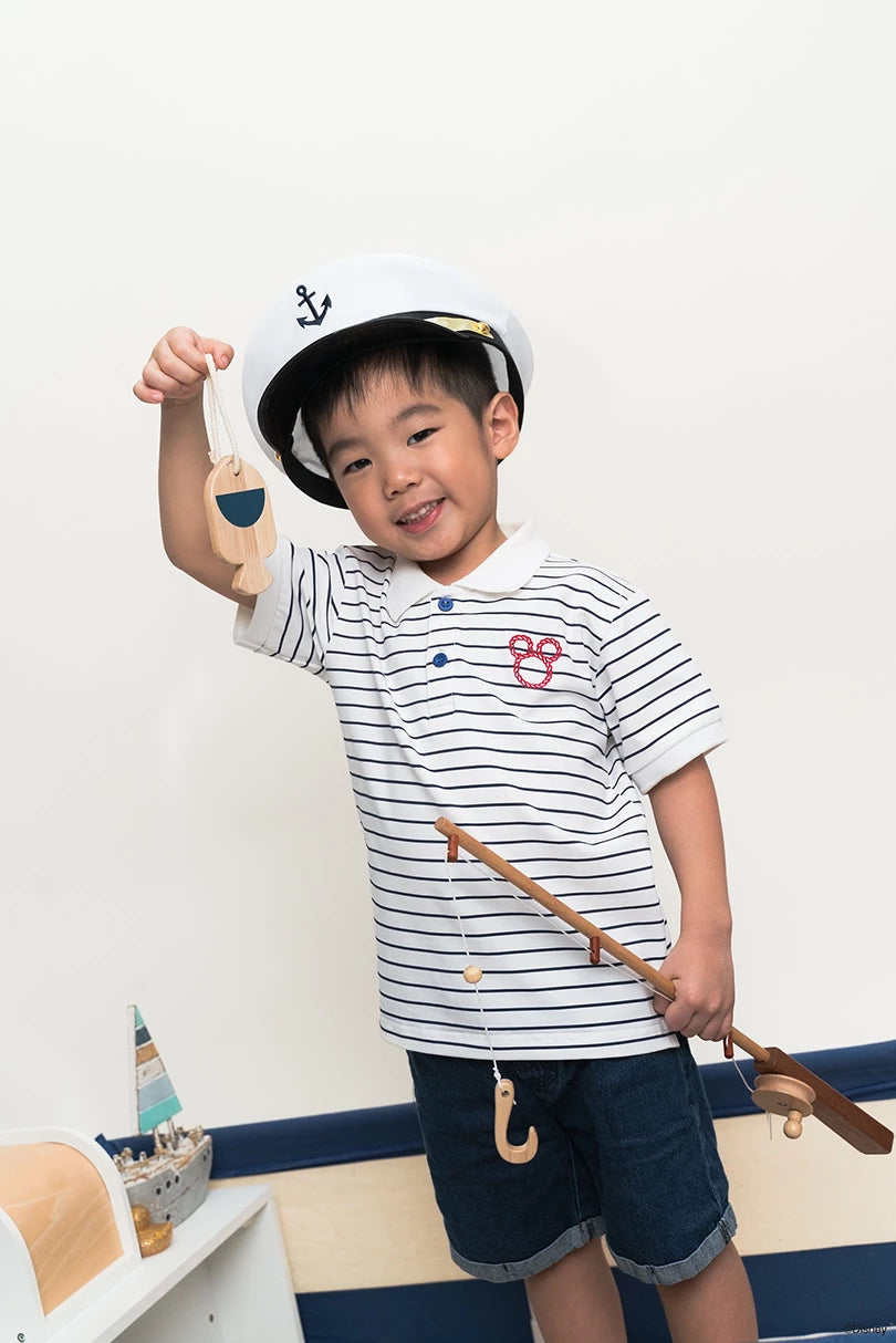 Child wearing polo tee in stripe sailor mickey and shorts, holding a toy boat and oar, with a white wall background.
