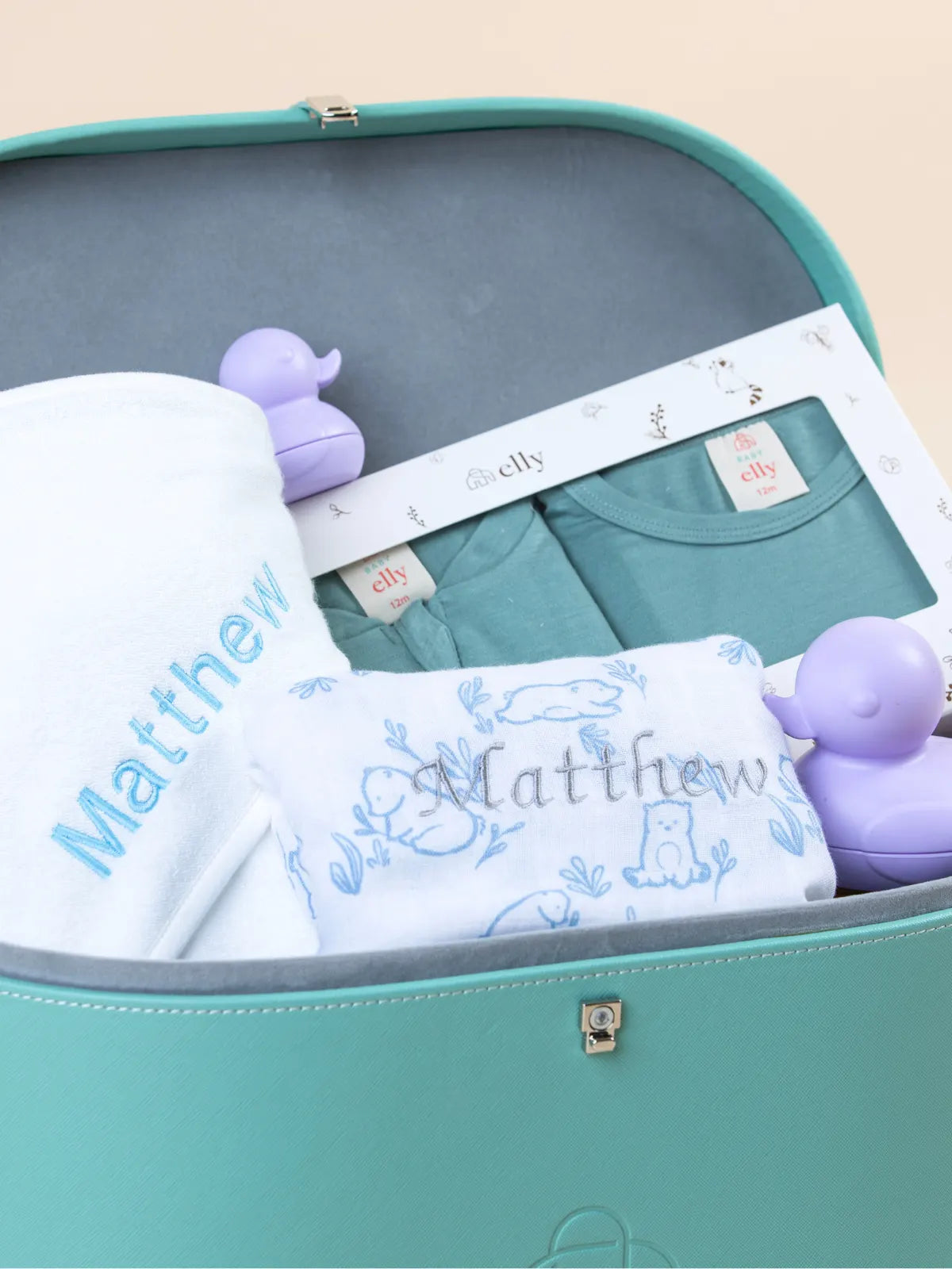 Close-up view inside the teal keepsake trunk showing a personalized white baby blanket embroidered with the name "Matthew" and matching teal apparel.