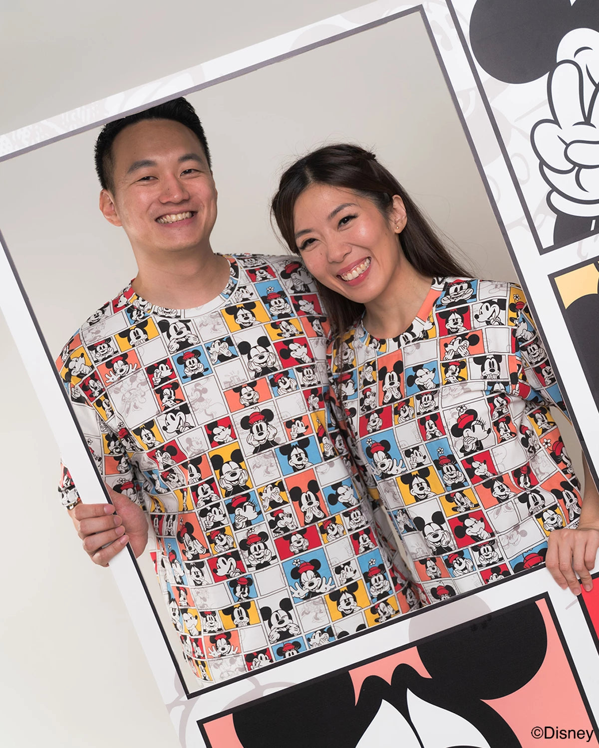 Two people wearing matching adult pop comic mickey tee standing next to a large poster with Mickey Mouse designs.