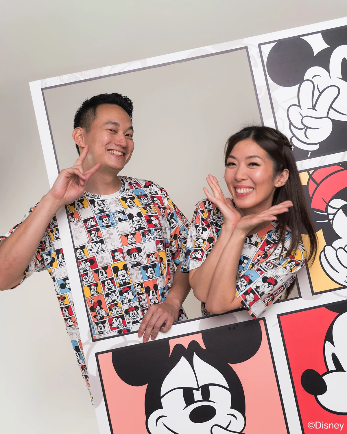 Two people wearing matching adult pop comic mickey tee standing next to a large poster with Mickey Mouse designs.