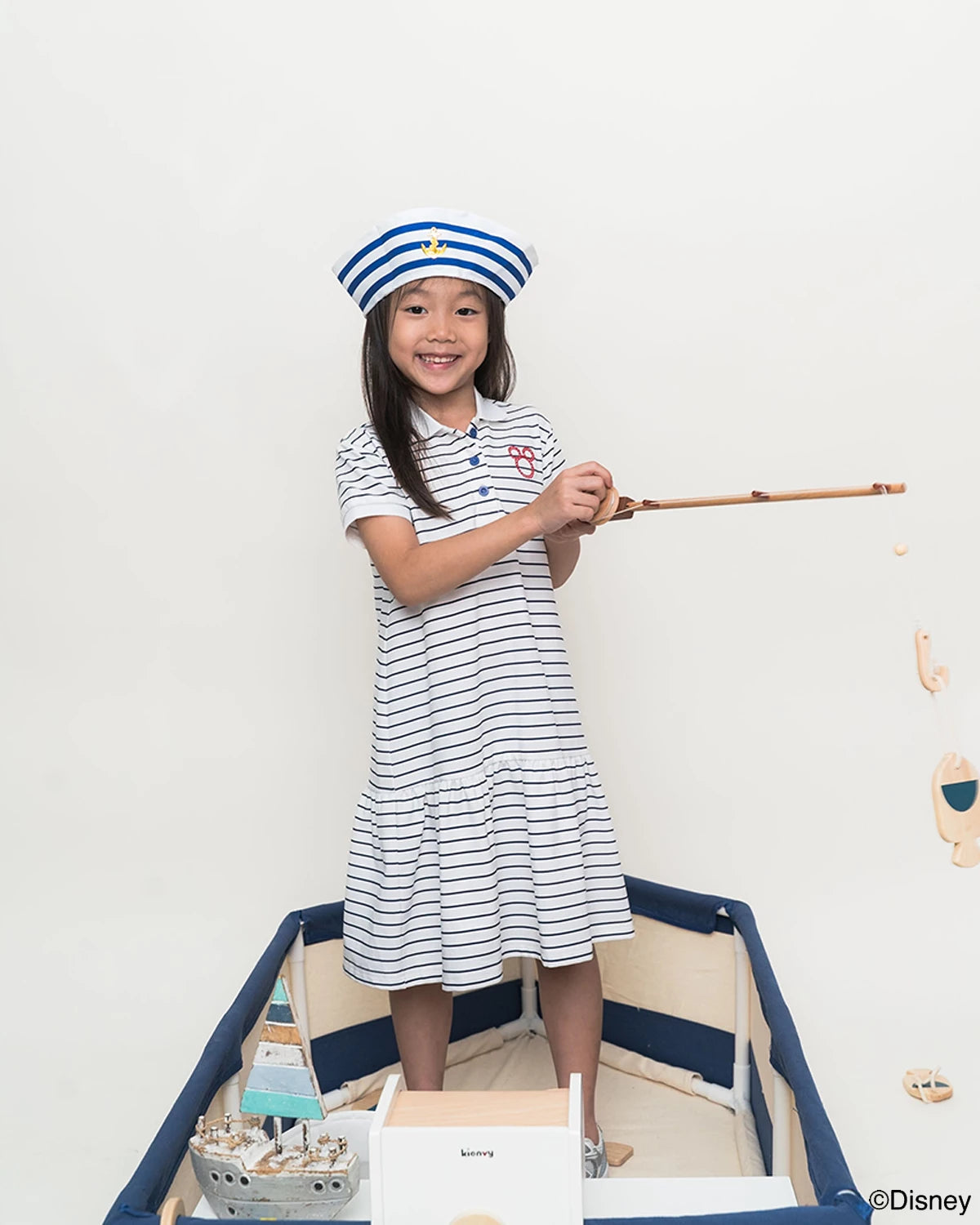 Child in sailor mickey stripes polo dress and sailor hat standing in a toy boat with a white background